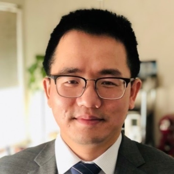Headshot of a person wearing a gray suit, white shirt, and blue-striped tie. They have short dark hair and are wearing black-framed glasses.