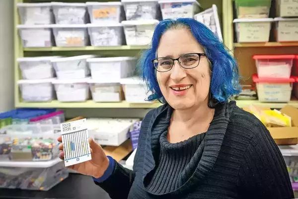 A person with bright blue hair, glasses, and a dark blue sweater smiles while holding a small printed card with a graph and QR code in front of shelves filled with containers.