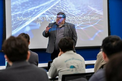 A man in a grey blazer and glasses gestures, presenting "Advanced 3D Stacking Technology for High Performance Computing" on a screen with a blue geometric background to an audience.